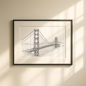 Puede incluir: Impresión enmarcada en negro del Golden Gate Bridge en San Francisco. La obra de arte es un dibujo detallado a pluma y tinta en blanco y negro del icónico puente colgante, sobre un fondo neutro. El puente se extiende sobre el agua con colinas al fondo.