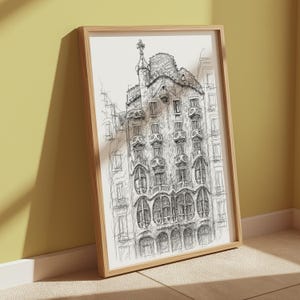 May include: A framed black and white architectural sketch of a building with ornate details, including arched windows and a decorative tower. The artwork is displayed in a light-colored wooden frame, leaning against a yellow wall.
