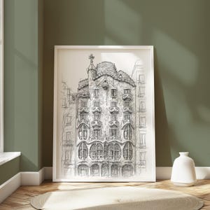 May include: A framed black and white architectural sketch of a building with ornate details, displayed against a green wall. The artwork is in a white frame and rests on a wooden floor with a small rug. A white vase is next to the frame.