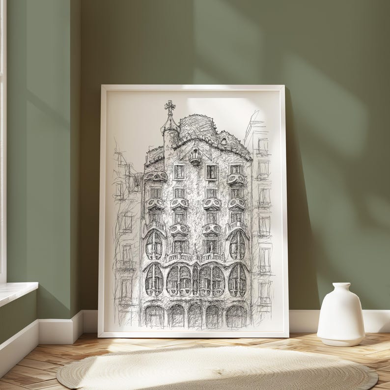 May include: A framed black and white architectural sketch of a building with ornate details, displayed against a green wall. The artwork is in a white frame and rests on a wooden floor with a small rug. A white vase is next to the frame.