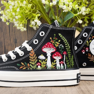 Puede incluir: Zapatillas Converse negras de caña alta con suelas blancas y un logotipo de estrella blanca. Las zapatillas están bordadas con un diseño floral que incluye setas rojas, hojas verdes y flores amarillas.