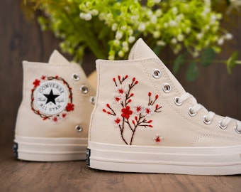 May include: A pair of off-white high-top Converse sneakers with embroidered red and white flowers. The sneakers have a white rubber sole and a black Converse All Star logo on the side.