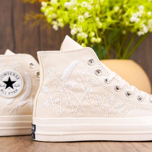 May include: A pair of off-white high-top Converse trainers with white embroidered floral designs. The trainers have black laces and a black Converse All-Star logo on the side.