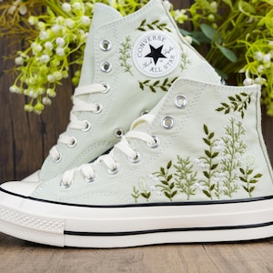 May include: Pair of high-top sneakers in a light green hue, featuring white laces and a white sole with black accents. The sneakers are adorned with embroidered floral designs in green and white, and the Converse All Star logo.