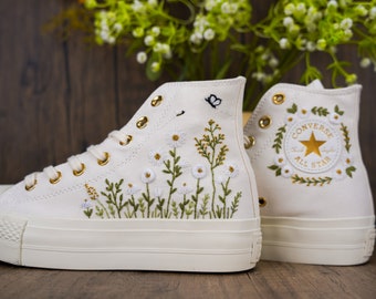 May include: White high-top sneakers with floral embroidery. The sneakers feature embroidered daisies, greenery, and a butterfly. Gold eyelets and the "Converse All Star" logo are visible on the back of the shoe.