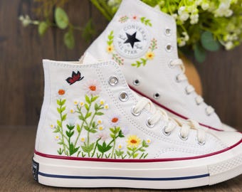 May include: White high-top canvas sneakers with embroidered flowers and a butterfly. The sneakers have a red trim around the sole and the words "Converse All Star" are embroidered on the side.