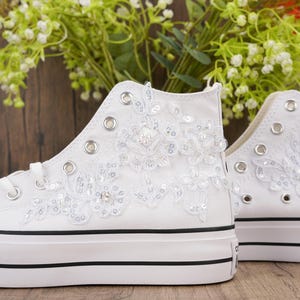 Puede incluir: Zapatillas altas blancas adornadas con encaje floral y lentejuelas. Los zapatos tienen una raya negra en la suela blanca y un logotipo de Converse All Star. Estas zapatillas son una opción elegante para ocasiones especiales.