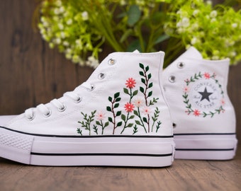 May include: White high-top canvas sneakers with floral embroidery. One shoe features a floral design, while the other has a Converse All Star logo with a floral border.