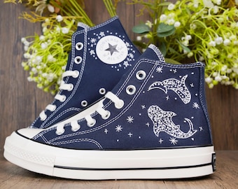 May include: A pair of navy blue high-top canvas sneakers featuring white lace-up closures, white rubber toe caps, and white soles. The outer side of the sneakers displays white hand-painted whale shark illustrations, small star motifs, and the Converse logo.