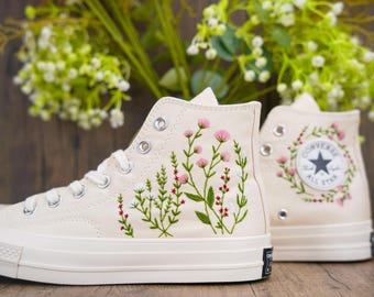 May include: A pair of white high-top canvas sneakers with embroidered floral designs. The sneakers have a black star logo on the side and a white rubber sole.