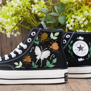 Könnte beinhalten: Schwarze High-Top-Sneaker mit weißen Schnürsenkeln und Sohlen. Die Sneaker haben gestickte Insekten und ein Schmetterlingsdesign. Das Converse All Star-Logo ist auf der Seite sichtbar. Die Schuhe stehen vor einem Holzgrund.