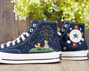 May include: Navy blue high-top sneakers with white laces and soles. The sneakers feature embroidered designs of a boy, fox, and stars on the sides. The Converse logo is visible on the back of one shoe.