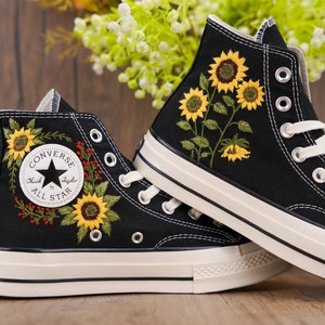 May include: Black high-top Converse sneakers with embroidered sunflowers and red berries. The Converse All Star logo is visible on the side of the shoe.
