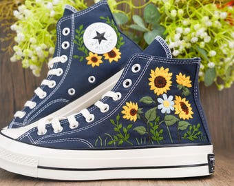 May include: Navy blue high-top sneakers with embroidered sunflowers and daisies. The sneakers have white laces, soles, and a classic Converse logo. The floral embroidery adds a unique, artistic touch to the footwear.