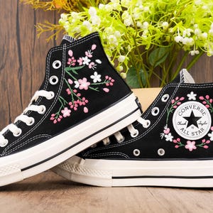 May include: Black high-top Converse sneakers with white soles and pink and white floral embroidery. The Converse logo is visible on the side of the shoe.