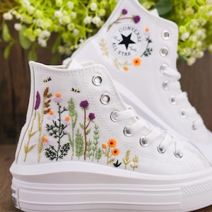 Può includere: Sneakers Converse bianche alte con fiori e foglie verdi ricamate. Le sneakers hanno una suola platform e un logo Converse All Star nero.