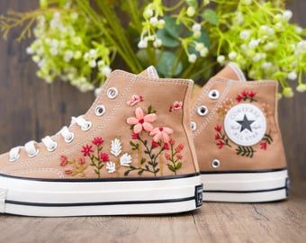May include: A pair of brown high-top Converse sneakers with white soles. The sneakers are embroidered with pink and red flowers and green leaves. The Converse logo is visible on the side of the sneakers.