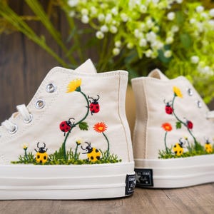 Könnte beinhalten: Ein Paar weiße High-Top-Canvas-Sneaker mit gestickten Blumen, Marienkäfern und gelben Käfern. Die Sneaker haben eine weiße Gummisohle.