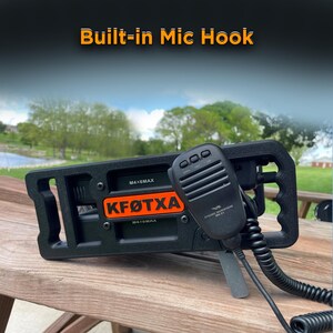 May include: A black radio transceiver mount featuring a built-in mic hook, an orange nameplate with the text KF&Oslash;TXA, and a black handheld dynamic microphone with a coiled cord. The device is secured with visible screws and includes M4X6MAX labels.