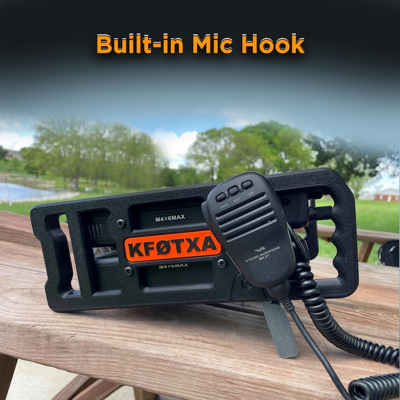 May include: A black radio transceiver mount featuring a built-in mic hook, an orange nameplate with the text KF&Oslash;TXA, and a black handheld dynamic microphone with a coiled cord. The device is secured with visible screws and includes M4X6MAX labels.