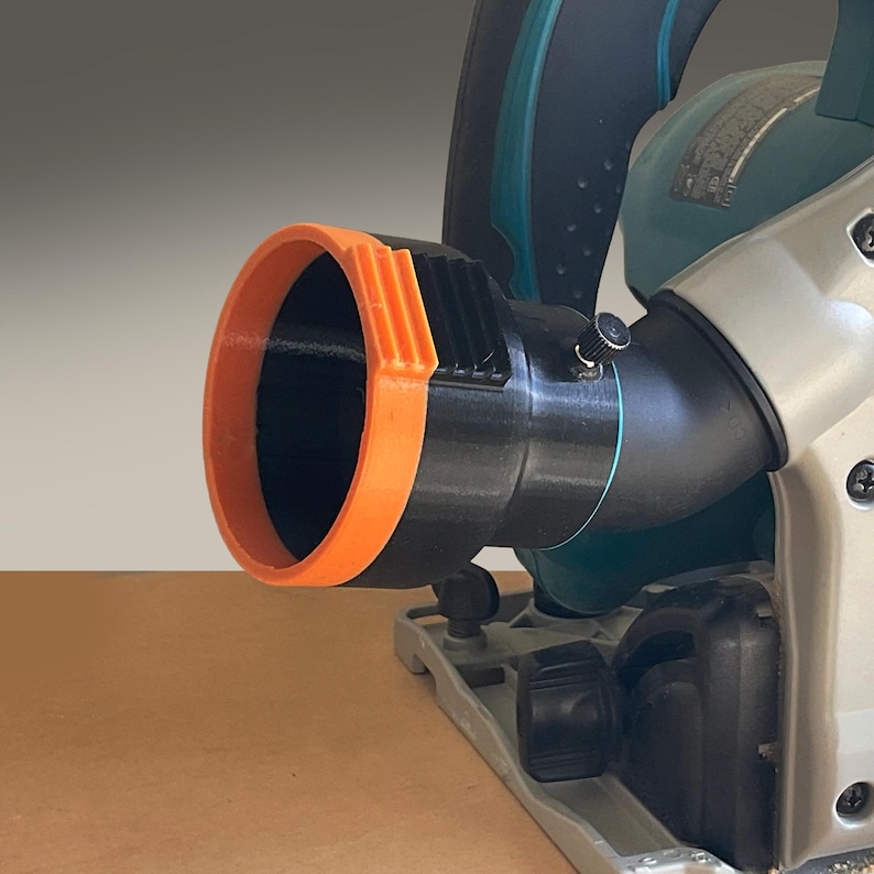 May include: Close-up of a black and orange circular saw dust collection attachment. The attachment is connected to a teal and silver circular saw. The orange ring has a series of vertical ridges.