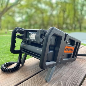 May include: A black amateur radio transceiver mounted in a protective frame with a coiled black microphone cord. The device features a large circular dial, a digital display, and an orange rectangular label with the text KF6TXA on the side.