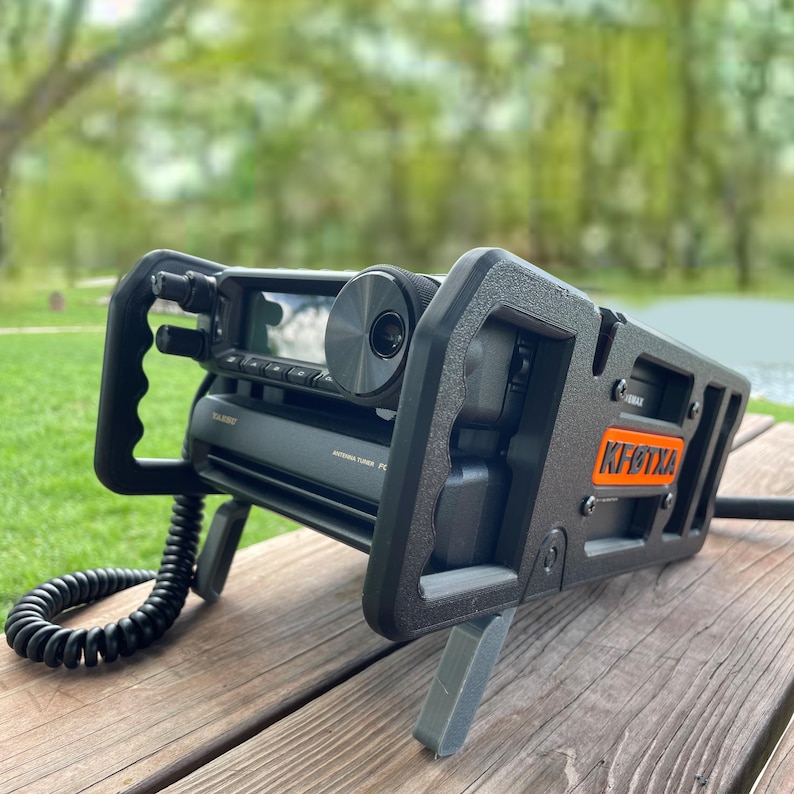 May include: A black amateur radio transceiver mounted in a protective frame with a coiled black microphone cord. The device features a large circular dial, a digital display, and an orange rectangular label with the text KF6TXA on the side.