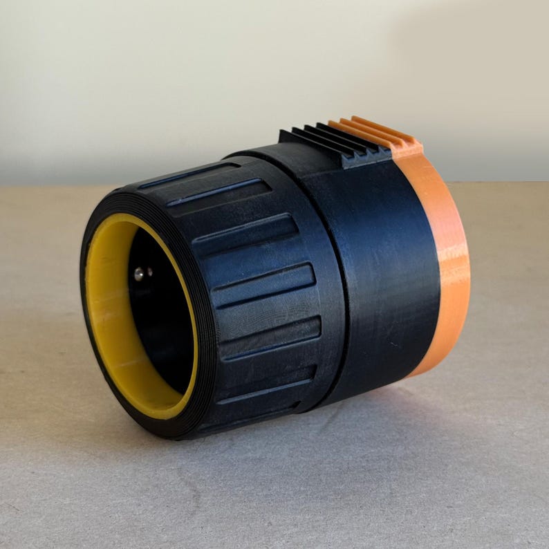 Puede incluir: Objeto cil&iacute;ndrico con cuerpo negro, anillo interior amarillo y detalles en naranja. El objeto tiene una secci&oacute;n estriada en la parte superior y parece estar hecho de pl&aacute;stico. Descansa sobre una superficie marr&oacute;n claro.