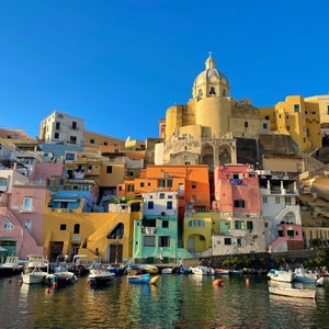 May include: A colorful village built on a hillside overlooking a harbor with boats docked in the water. The buildings are painted in shades of yellow, orange, pink, blue, and green. A white church with a dome sits atop the hill.