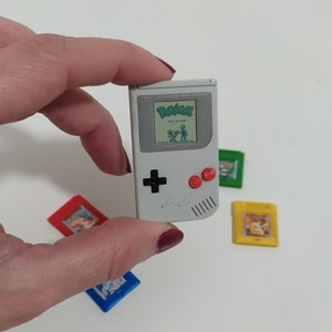 May include: A miniature gray Game Boy with a screen displaying "Pokémon" text and graphics, held by a hand. Several colorful game cartridges in red, blue, green, and yellow are scattered around the console.