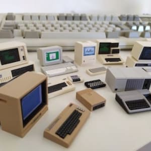 Puede incluir: Una colección de ordenadores vintage en miniatura, incluyendo un Apple IIe beige, un IBM PC blanco y un Commodore 64 gris. Los ordenadores están dispuestos sobre una superficie blanca con un teclado en el fondo.
