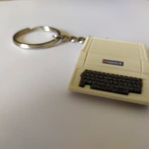 Könnte beinhalten: Ein weißer Plastik-Schlüsselanhänger in Form eines alten Computers mit einer schwarzen Tastatur und dem Wort "COMMODORE" auf der Oberseite.