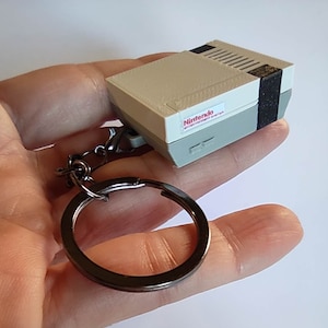 Op de afbeelding: Een grijze en zwarte Nintendo Entertainment System sleutelhanger met een zilveren sleutelring. De sleutelhanger is een miniatuurreplica van de klassieke videogameconsole.