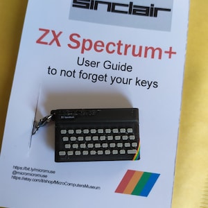 Op de afbeelding: Een zwarte ZX Spectrum+ sleutelhanger met een regenboogstreep, op een witte kaart. De kaart toont het Sinclair-logo en de tekst "ZX Spectrum+ User Guide to not forget your keys."