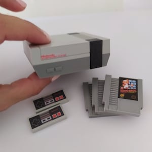 May include: A miniature Nintendo Entertainment System (NES) console with two controllers and four game cartridges. The console is gray and white with a black stripe. The cartridges are gray with black labels. The controllers are gray with red buttons.