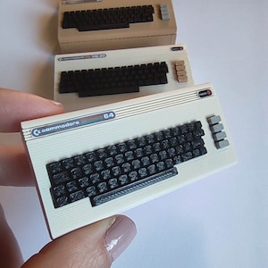Könnte beinhalten: Ein Set aus drei Miniatur-Vintage-Computer-Tastaturen. Die oberste Tastatur ist schwarz mit weißen Tasten. Die mittlere Tastatur ist beige mit schwarzen Tasten und dem Text "VIC 20". Die unterste Tastatur ist weiß mit schwarzen Tasten und dem Text "commodore 64".