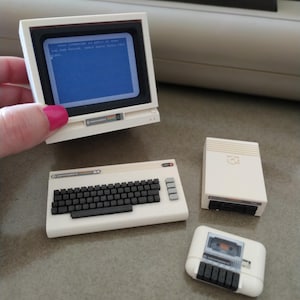 Può includere: Un set di computer Commodore 64 bianco in miniatura, che include un monitor che mostra testo, una tastiera, un'unità disco e un lettore di cassette. Il monitor è tenuto da una persona. La tastiera ha tasti neri.
