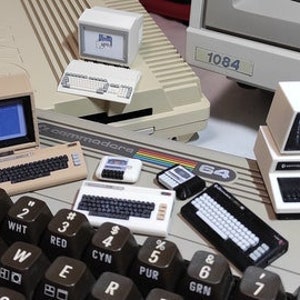 Op de afbeelding: Een verzameling miniatuur vintage computers, waaronder een Commodore 64, een Apple II en een TRS-80. De computers zijn gerangschikt op een zwart toetsenbord met toetsen die zijn gelabeld met nummers en kleuren.