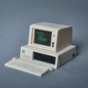 Puede incluir: Un ordenador de sobremesa blanco de estilo vintage con monitor, teclado y unidad de sistema. El monitor muestra texto verde sobre un fondo negro.