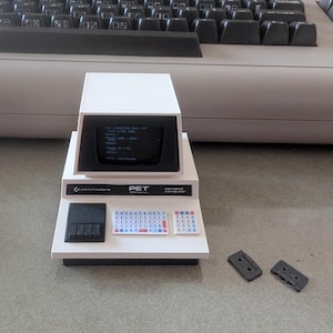 Puede incluir: Un modelo de computadora personal Commodore PET serie 2001 de color blanco con una pantalla negra con texto, un teclado pequeño, un teclado numérico y un área de casetera negra. Dos pequeños componentes rectangulares negros se sitúan a la derecha.
