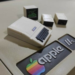 May include: A miniature replica of an Apple IIc computer with a beige plastic case, a black keyboard, and a green screen. Three additional beige plastic boxes are included, likely representing floppy disk drives.