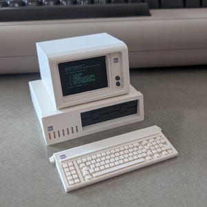 Può includere: Un modello di computer in miniatura color bianco sporco con un monitor con display a schermo verde, un'unità centrale con uno slot per unità disco nero e una tastiera abbinata. Il logo IBM è visibile sul monitor, sulla CPU e sulla tastiera.