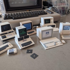 May include: A collection of miniature vintage computer models including a Commodore PET, an Apple Macintosh with a hello screen, and an Amiga, arranged alongside a full-sized vintage computer keyboard and monitor, with small black floppy disk accessories.