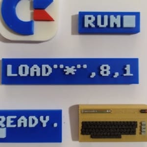 May include: A set of five blue and white plastic buttons with retro computer-themed text. The buttons read "RUN", "LOAD ""*", 8, 1", "READY.", and a blue and red C logo.