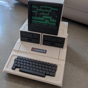 Può includere: Un computer Apple II bianco con una tastiera nera e due unità floppy disk. Sopra le unità floppy disk c'è una piccola replica in 3D stampata in nero di un computer Apple II con uno schermo verde che mostra un videogioco. Il testo sullo schermo dice "SCORE 14410  REMAIN 03  LEVEL 001".