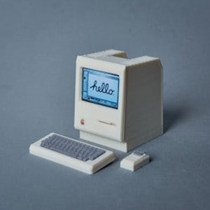 Puede incluir: Una réplica en miniatura de plástico blanco de una computadora Apple Macintosh con un teclado y un ratón. La pantalla de la computadora muestra la palabra "hello" en una fuente de escritura a mano.
