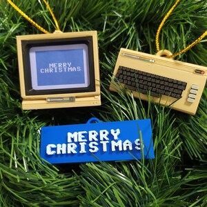 Peut inclure: Deux décorations de Noël en forme d'ordinateurs vintage. L'un est un Commodore 64 beige avec un clavier noir et l'autre est un moniteur Commodore beige avec un écran bleu qui affiche "Joyeux Noël". Une décoration bleue avec du texte blanc qui dit "Joyeux Noël" est suspendue sous les ordinateurs.