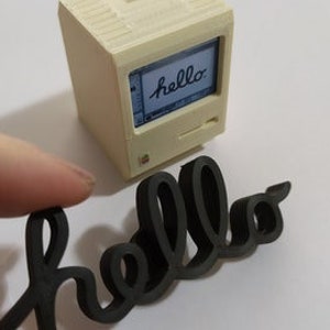 Op de afbeelding: Een miniatuur witte computer met een zwart scherm waarop het woord "hello" wordt weergegeven. Op de voorgrond bevindt zich een zwarte houten uitsnijding van het woord "hello".
