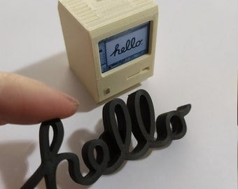 May include: A miniature white computer with a black screen displaying the word "hello". A black wooden cutout of the word "hello" is in the foreground.