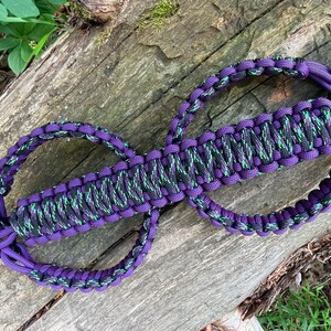 May include: A paracord handle featuring a braided design in solid purple and a black cord with green flecks. The handle is constructed with a central woven section and two circular loops on either end, finished with a black metal shackle attachment.